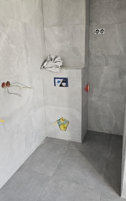 Gefliestes Badezimmer im Rohbau mit großformatigen, hellgrauen Wand- und Bodenfliesen sowie sichtbaren Wasser- und Elektroanschlüssen.