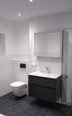 Badezimmer mit schwarzen Bodenfliesen, weißen Wandfliesen, mit Toilette, Spiegelschrank und Wasserhahn mit schwarzem Unterschrank.