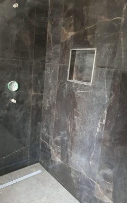 Dusche im Rohbau mit dunklen Wandfliesen im Marmormuster sowie sichtbaren Wasseranschlüssen und Wandnische als Ablage.