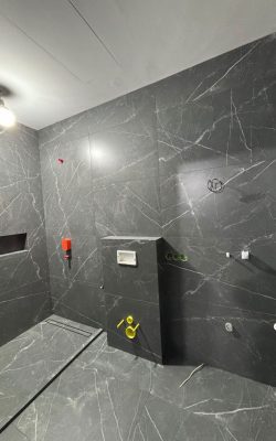 Badezimmer im Rohbau mit dunkelgrauen Wandfliesen im Marmormuster sowie sichtbaren Wasser- und Elektroanschlüssen.