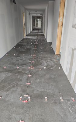 Verlegter Boden in einem langen Flur mit großen grauen Fliesen, die mithilfe von roten Nivelliersystemen und Abstandshaltern fixiert werden.