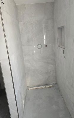Dusche im Rohbau mit hellgrauen Wandfliesen sowie sichtbaren Wasseranschlüssen und Wandnische als Ablage.
