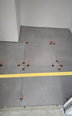 Verlegter Boden mit großen grauen Fliesen, die mithilfe von roten Nivelliersystemen und Abstandshaltern fixiert werden.