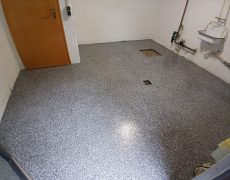 Graumelierter Boden in Raum mit Waschbecken und Wandleitungen.