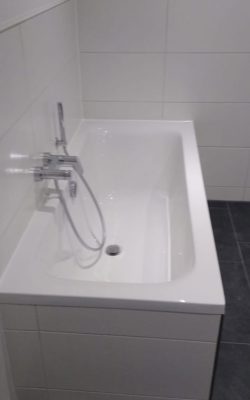 Badewanne auf schwarzen Bodenfliesen und vor weißen Wandfliesen.